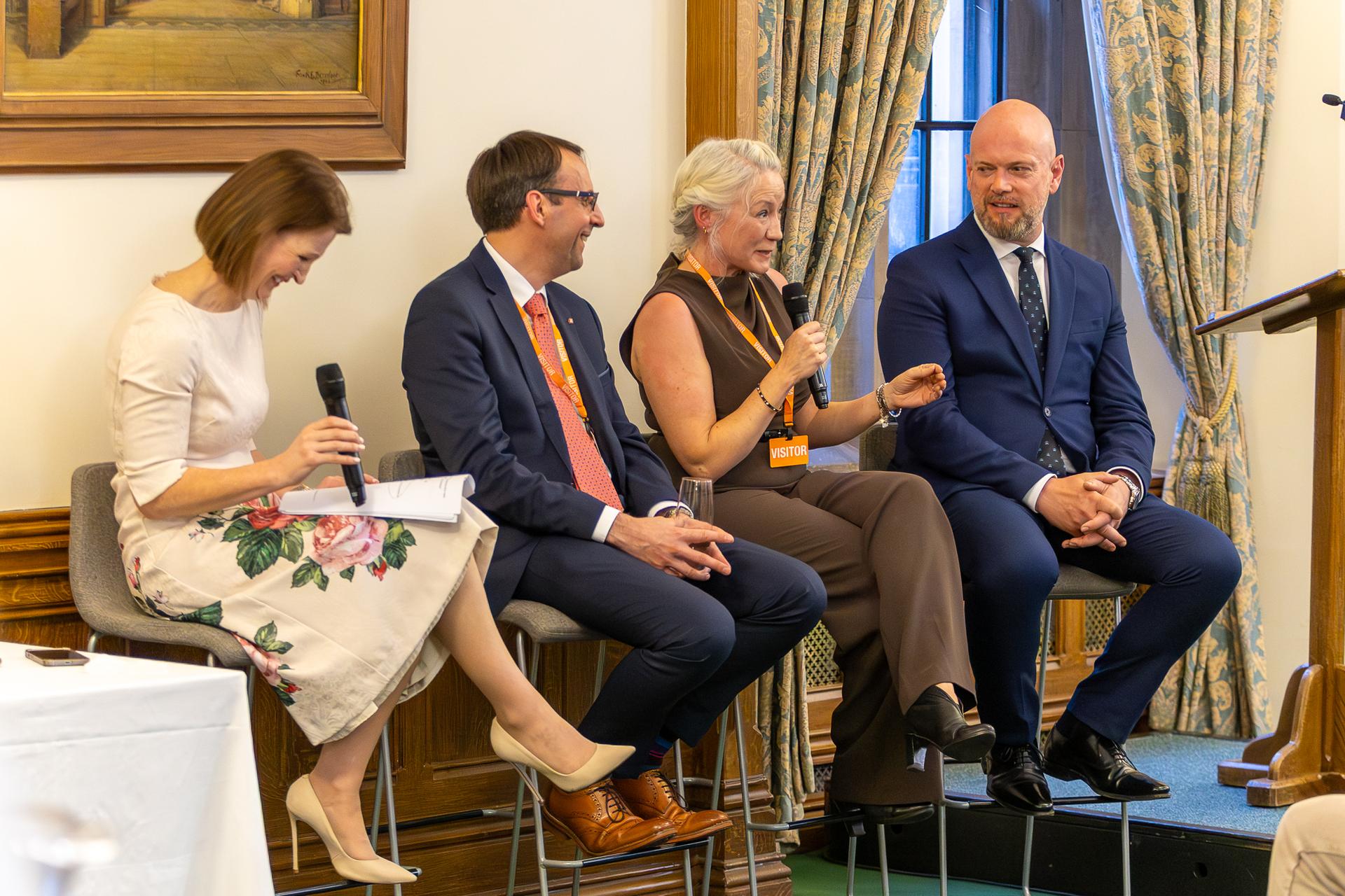 Four speakers sit on a stage panel during a discussion event in an ornate room with wood-panelled walls, a framed painting and patterned curtains. Three participants hold handheld microphones while seated on high chairs beside a lectern, engaging in a moderated conversation before an audience.